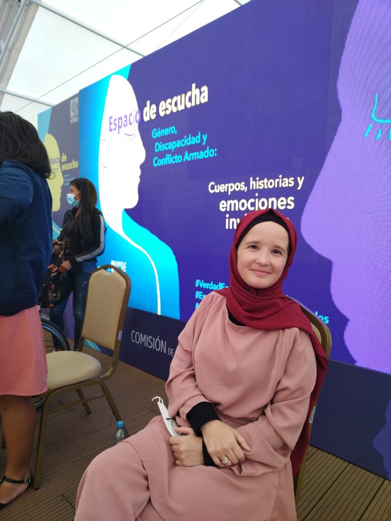 Sabrina PT, integrante de Polimorfas, mujer de tez blanca, lleva un vestido color rosa y un hijab rojo. Está sentada y en el fondo se proyecta una imagen con rostros de mujeres azules con los siguientes textos: “Espacio de escucha. Género, Discapacidad y Conflicto Armado “ y “Cuerpos, historias y emociones invisibles”