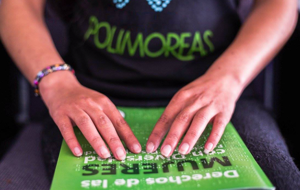 Una mujer que lleva una camiseta de polimorfas tiene una cartilla verde sobre sus piernas. Esta cartilla tiene el título “derechos de las mujeres con diversidad”. La mujer tiene las yemas de sus dedos sobre la cartilla tocando en relieve el texto en sistema Braille.