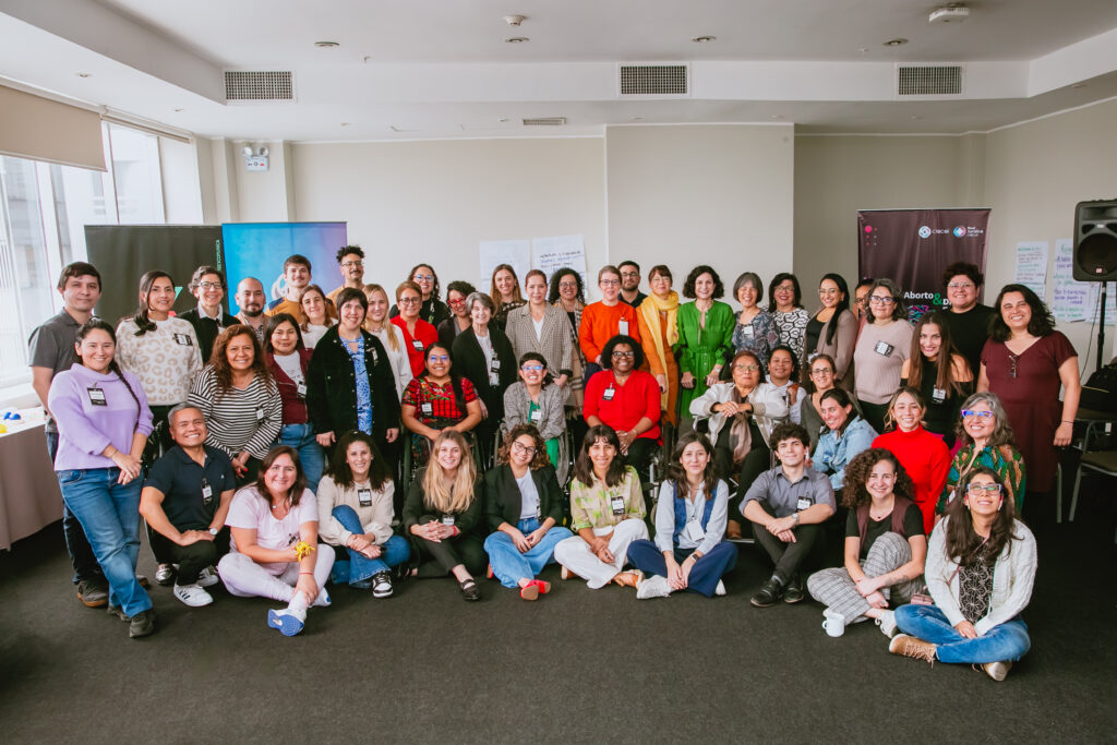 Fotografía con 51 personas, en su mayoría mujeres, con distintas identidades etnico raciales y algunas de ellas con discapacidad. Las personas están mirando a la cámara y están reunidas en semicírculo en filas de diferente altura. Al fondo se ven pendones y algunos pliegos de papel de trabajo.
