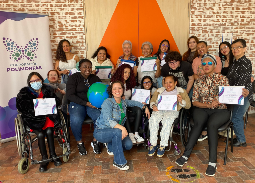 18 mujeres con diversas discapacidades participantes del evento “encuentro de mujeres disca, diversas y libertas” sosteniendo sus certificados de participación. Las mujeres están algunas de pie y otras sentadas en el piso o en sus sillas de ruedas.