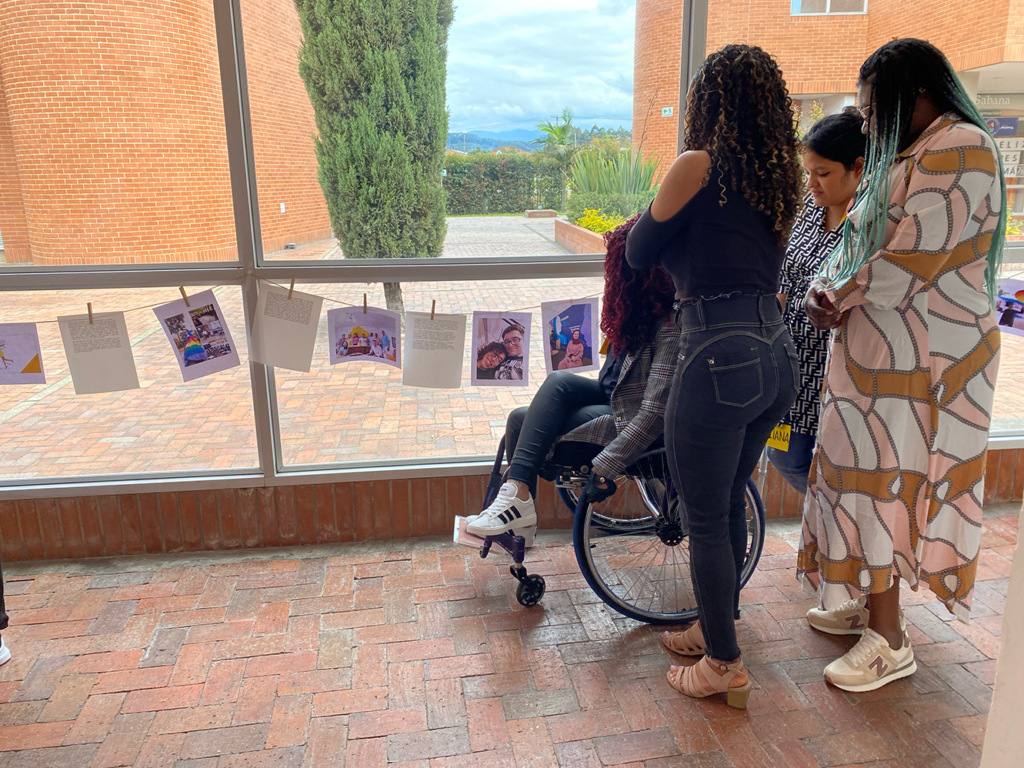 4 mujeres con discapacidad están observando diferentes fotografías que están colgadas sobre un cristal de la actividad "El tendedero". 3 de ellas están de pie, mientras que una de ellas está sentada en una sillas de ruedas