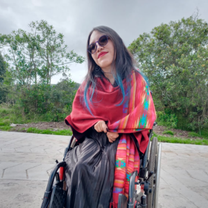 Mujer mestiza de cabello negro con puntas azules lacio, lleva unas gafas oscuras. Está sentada sobre una silla de ruedas y lleva un chal rojo con un diseño tejido en distintos colores. En el fondo se ve un bosque.