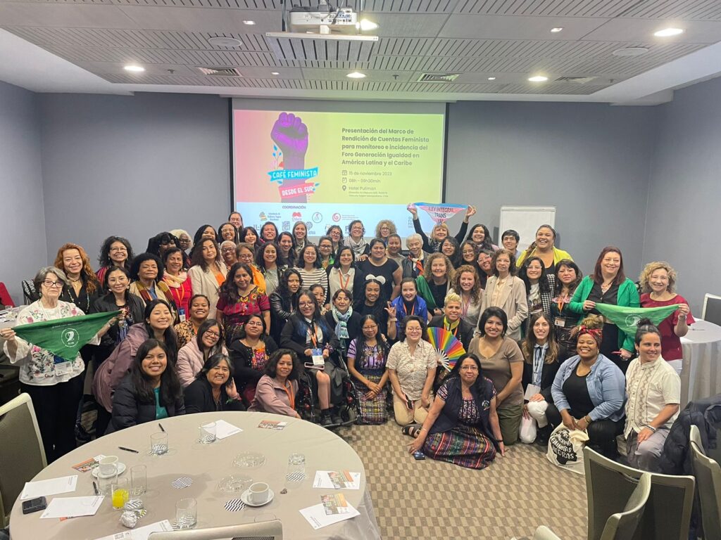 Grupo de mujeres participantes del MFRC Consenso de montevideo reunidas ondeando diversas pañoletas: verdes, de la bandera trans y de la bandera LGTBIQ+. En el fondo se proyecta una presentación que dice “ Presentación del Marco de Rendición de Cuentas Feminista para monitoreo e incidencia del Foro Generación Igualdad en América Latina y el Caribe” Al lado izquierdo aparece una mano con un texto que dice “Cafe feminista desde el sur”