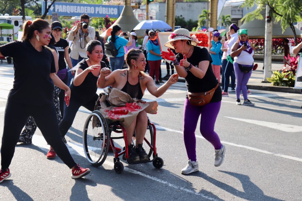 Mujeres con discapacidad en una marcha. En primer plano se ven 4 mujeres con discapacidad vestidas de negro en un performance en la que 3 de ellas de pie rodean a otra mujer usuaria de silla de ruedas. Cada una de las mujeres de pie tienen diferentes expresiones en su rostro mientras ven a la mujer usuaria de silla de ruedas que está empuñando sus manos y sonríe. Al fondo se ven otras personas participantes de la marcha.