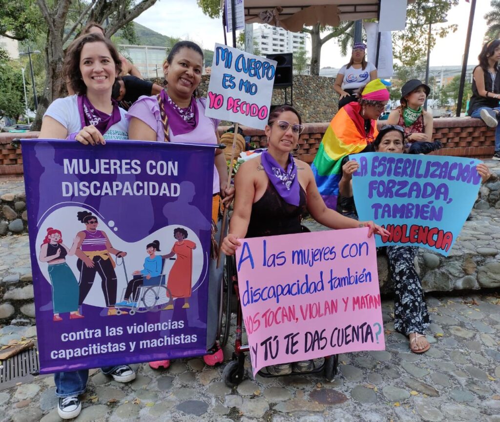 Cuatro mujeres con pañoletas moradas sostienen distintos carteles. De izquierda a derecha la primera mujer sostiene un cartel que dice “mujeres con discapacidad contra las violencias capacitistas y machistas”, la segunda mejor sostiene un cartel que dice “mi cuerpo es mío yo decido”, la tercera mujer, usuaria de silla de ruedas, sostiene un cartel que dice “ a las mujeres con discapacidad también nos tocan, violan y matan ¿ y tu te das cuenta?”, la última mujer sostiene un cartel que dice “la esterilización forzada también es violencia”.