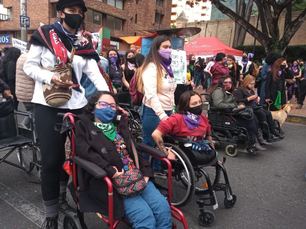 En primer plano se muestran en la calle cuatro mujeres en silla de ruedas que asisten a una marcha del 8M. Una de ellas es Bubulina Moreno, fundadora de Polimorfas. Detrás de ellas se ven sus acompañantes y las demás personas participantes de la marcha.