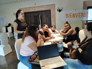 Cinco mujeres están sentadas en un mesa rectangular, una de ellas está comunicándose en lengua de señas y con apoyo de su intérprete. Las otras 4 mujeres prestan atención a su conversación