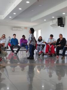 Bubulina Moreno, una mujer con una talla por debajo del promedio vestida con una camiseta blanca con flores azules, está en el centro de una recinto con mujeres con discapacidad que están a su alrededor en un círculo sentadas prestando atención a su exposición.