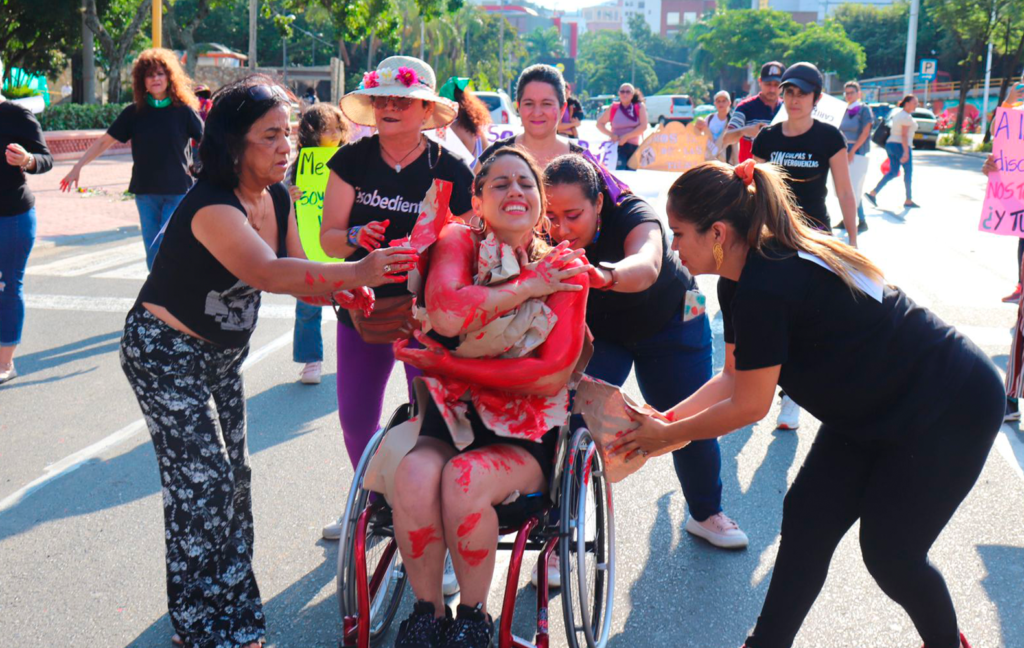 Mujeres con discapacidad en una marcha. En primer plano se ven 4 mujeres con discapacidad vestidas de negro en un performance en la que 3 de ellas de pie rodean a otra mujer usuaria de silla de ruedas. Cada una de las mujeres de pie están colocando un papel lleno de pintura roja a la usuaria de silla de ruedas, quien tiene una expresión de dolor y angustia. Al fondo se ven otras personas participantes de la marcha.