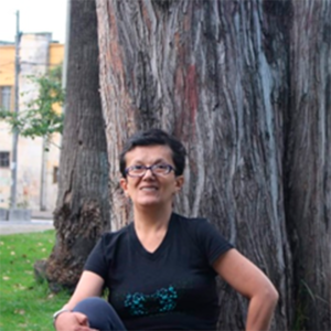 Mujer de piel morena con cabello corto negro y ojos color negro. Usa anteojos. Está sentada frente a un árbol vistiendo una camiseta negra con el logo de Polimorfas.