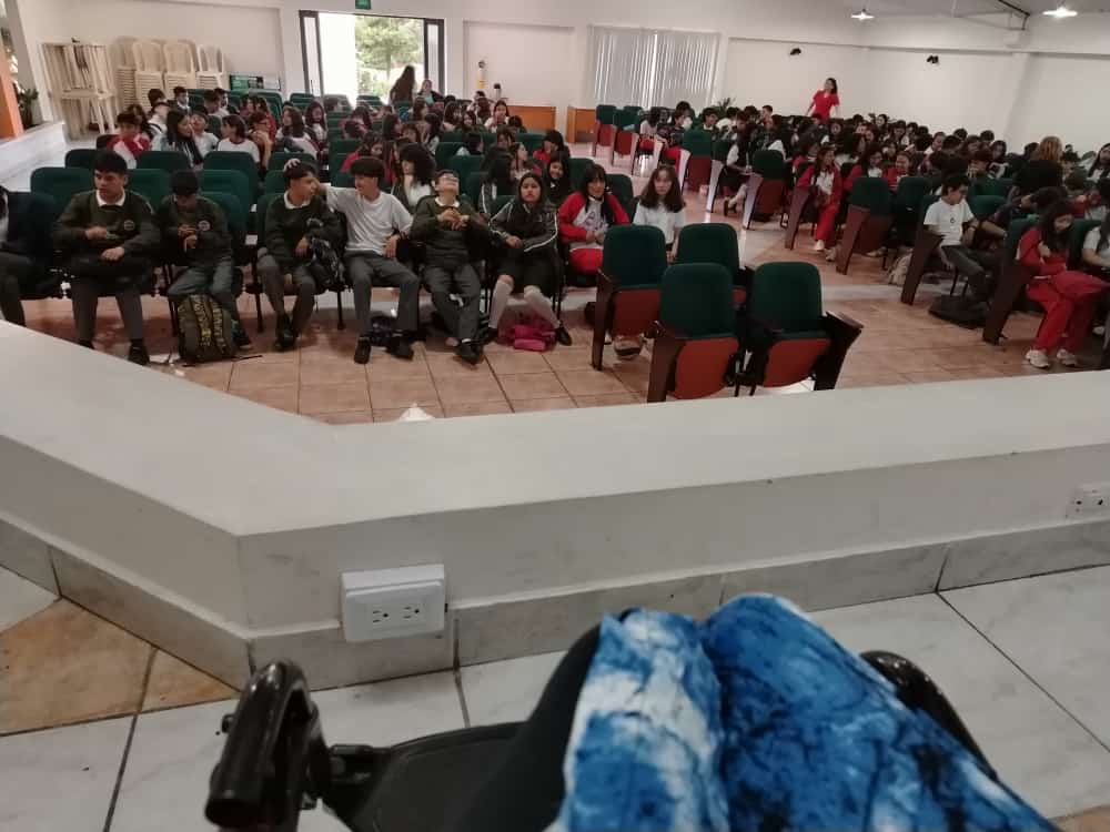 Fotografía tomada desde el ángulo de una persona usuaria de silla de ruedas que está en la tarima. A la distancia se ven las sillas de un auditorio donde están sentados niños, niñas y adolescentes. 