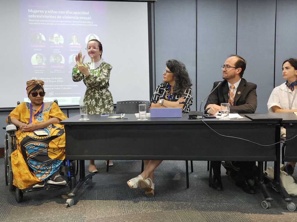 Cuatro mujeres y un hombre en el panel del evento  “Mujeres y niñas con discapacidad sobrevivientes de violencia sexual”. De izquierda a derecha aparece una mujer afro en silla de ruedas vestida con una bata tradicional y turbante amarillo, Sabrina mujer Sorda señando su intervención al publico vestida con un vestido verde, una mujer trigueña vestida con una blusa negro que dirige su mirada a la izquierda, un hombre trigueño en un vestido de paño, y una mujer trigueña vestida de camisa blanca y pañoleta azul.