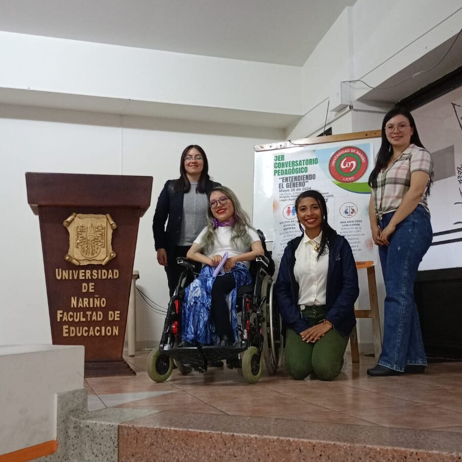 Sobre una tarima están 4 mujeres al lado de un pedestal. Dos de ellas están de pie, una está arrodillada en el suelo y una de ellas está sentada en su silla de ruedas. Detrás de ellas hay un tablero con una poster que dice “3er conversatorio pedagógico “Entendiendo el género”.