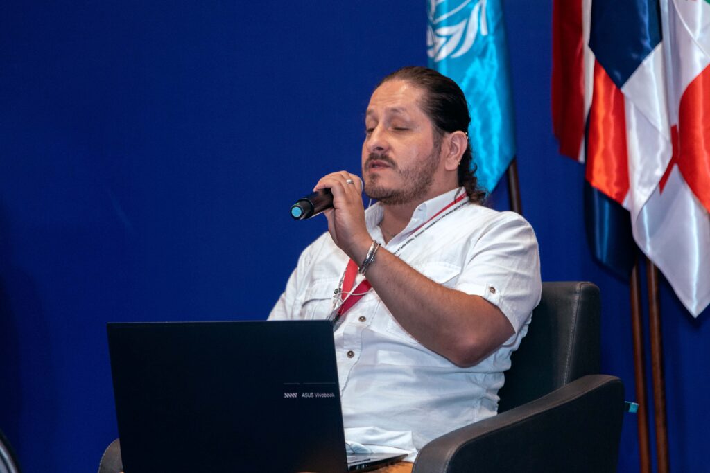 Primer plano de la intervención de Anderson Henao, Director para la Garantía de los Derechos de la Personas con Discapacidad del Ministerio de Igualdad y Equidad de Colombia. Hombre ciego de cabello crespo recogido, va vestido de camisa blanca, lleva un micrófono en su mano y un portatil negro en sus piernas.