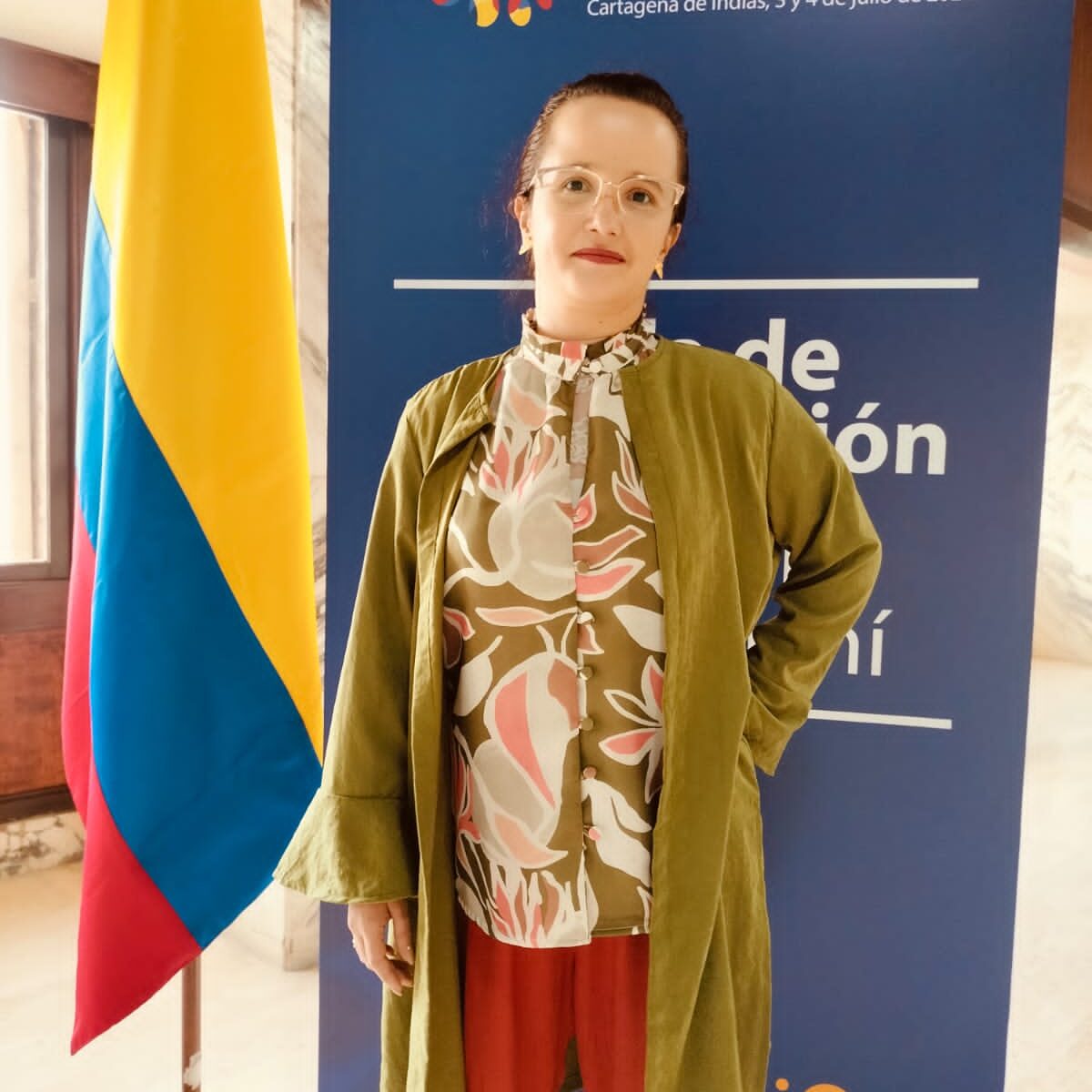 Primer plano de Sabrina P.T., Directora de Corporación Polimorfas. Mujer de tez blanca, cabello recogido en trenza, viste una camisa verde de flores rosas, pantalón naranja y chaqueta verde. Detrás de ella aparece un banner que dice V Conferencia Regional sobre Población y Desarrollo de América Latina y el Caribe. Cartagena de Indias 3 y 4 de Julio de 2024. Al lado izquierdo se ve la bandera de Colombia